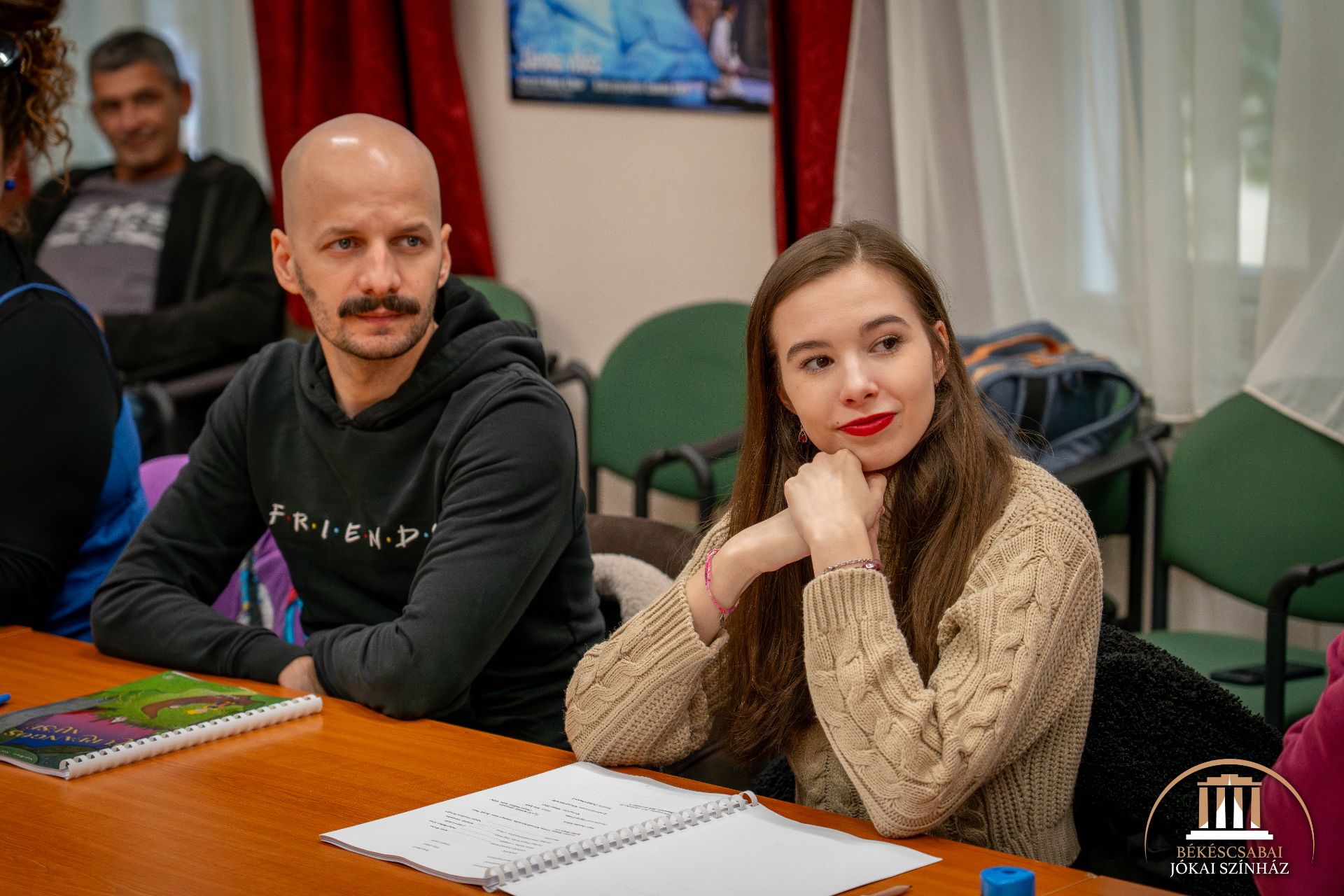 Egy fekete kapucnis férfi és egy bézs színű pulóvert viselő nő ül egy asztalnál, kezükben forgatókönyvekkel és jegyzetfüzetekkel, figyelmesen néznek. A közelben mások is ülnek, a sarokban a Békscsabai Jókai Színház logója látható.
