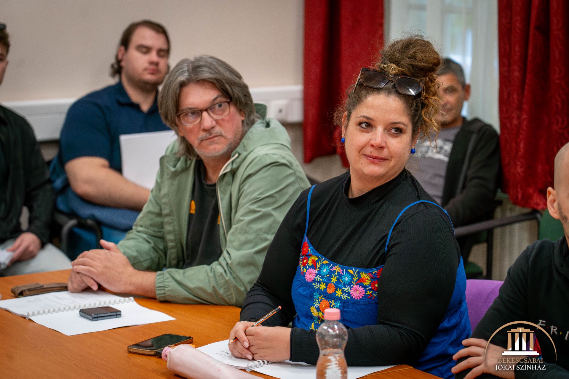 Egy megbeszélés vagy workshop során többen ülnek egy asztalnál, előttük füzetekkel és tollakkal. Egy kék virágos ruhás nő és egy zöld kabátos férfi van a fókuszban, mindketten figyelmesen hallgatnak.