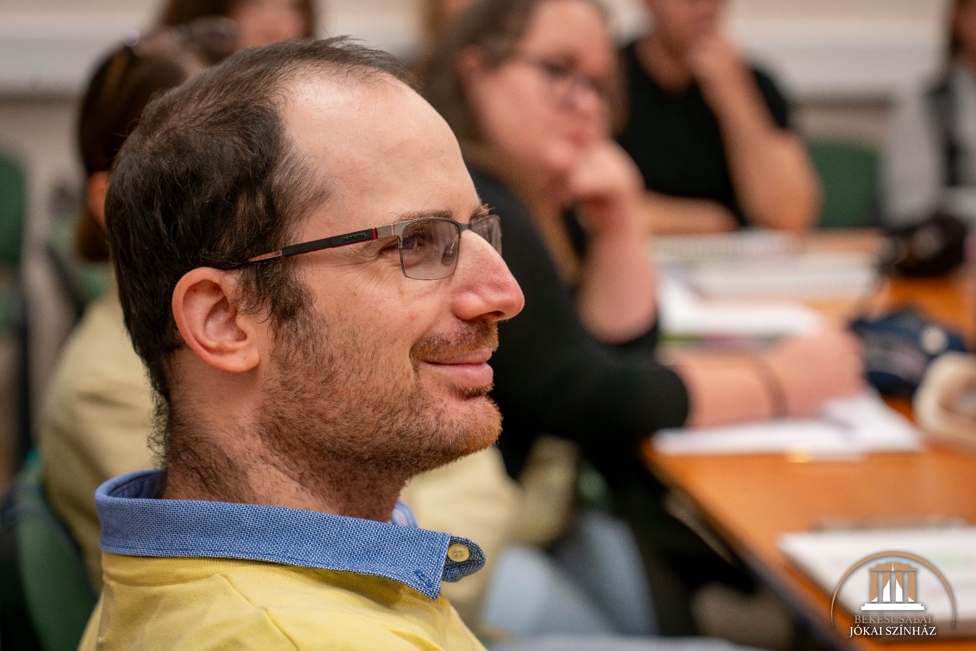 Egy szemüveges, sárga inges férfi ül egy asztalnál, mosolyog, a háttérben elmosódottan más emberek, ami tanteremre vagy tárgyalási helyszínre utal.