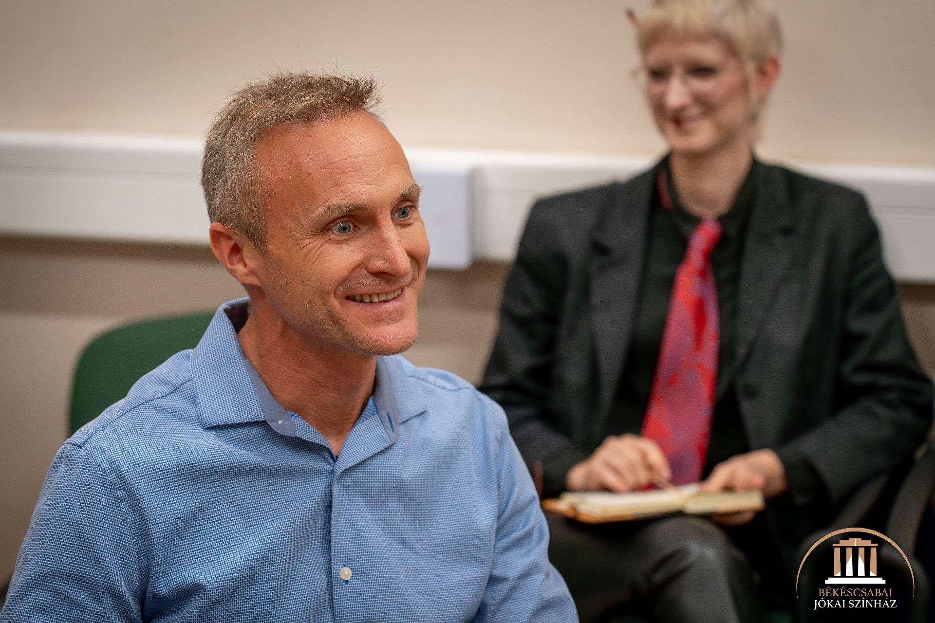 Az előtérben egy kék inges, mosolygós férfi ül, míg a háttérben egy öltönyös, szemüveges, piros nyakkendős ember ül, kezében egy notebookkal. A jobb alsó sarokban a Békéscsabai Jókai Színház logója látható.
