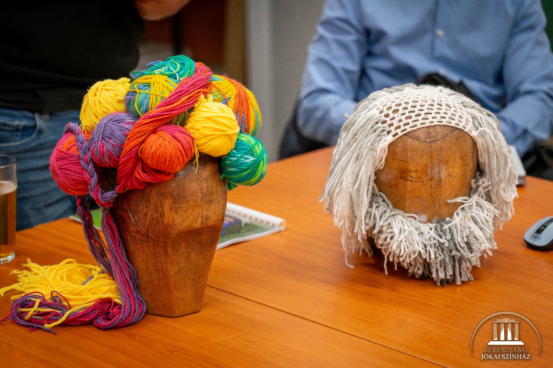 Két fából készült próbababa fej egy asztalon: az egyikre színes fonalból készült haj, a másikra fehér horgolt paróka és szakáll van erősítve. A háttérben egy pohár víz és egy jegyzetfüzet.