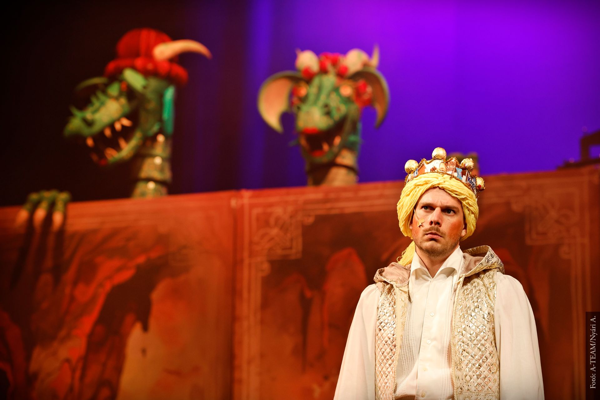 Színpadon egy koronás férfi áll komoly arckifejezéssel, mögötte két zöld, többfejű sárkánybábu emelkedik ki a díszlet mögül, színes, lila megvilágításban.