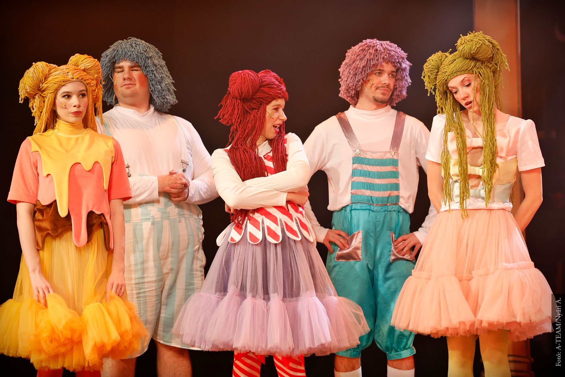 Öt színész áll egymás mellett a színpadon színes, játékos jelmezekben és parókákban. Különböző pózokat vesznek fel, miközben a háttér meleg fényben úszik.