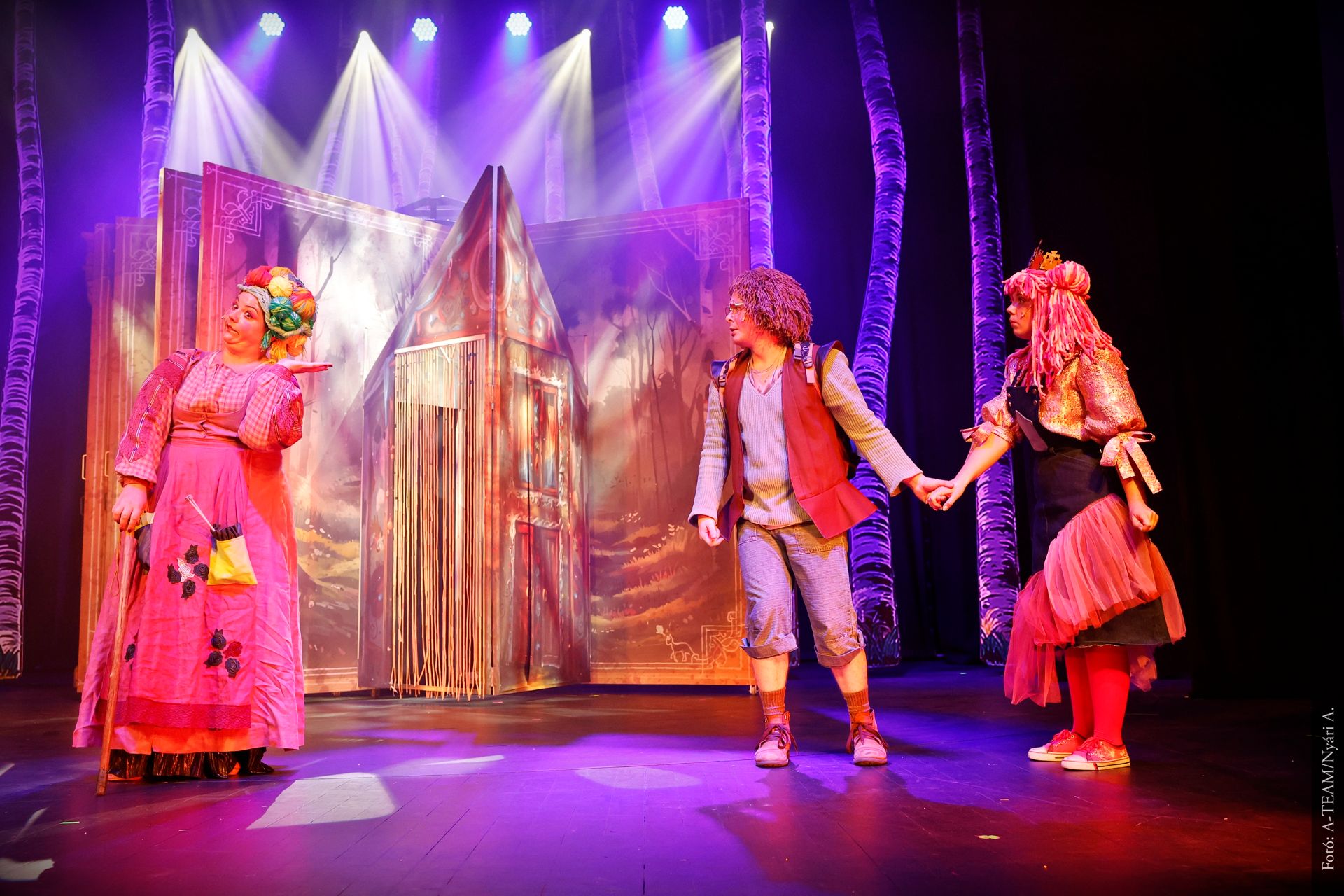 Három színész áll a színpadon, színes jelmezekben. A középen álló férfi kézen fogja a rózsaszín parókás nőt, miközben egy másik, élénk ruhás nő alakhoz beszélnek. A háttérben ház alakú díszlet és erdei háttér látható.