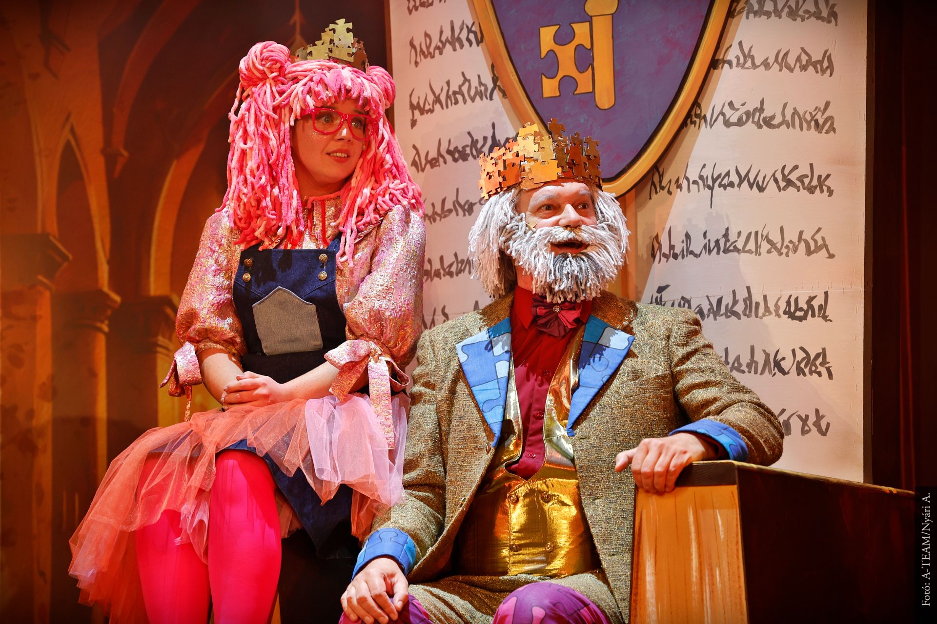 Színpadon egy koronás férfi és egy rózsaszín parókás nő ülnek egymás mellett. A háttérben díszes pajzs és aranybarna díszlet látható, a jelenet vidám hangulatú.
