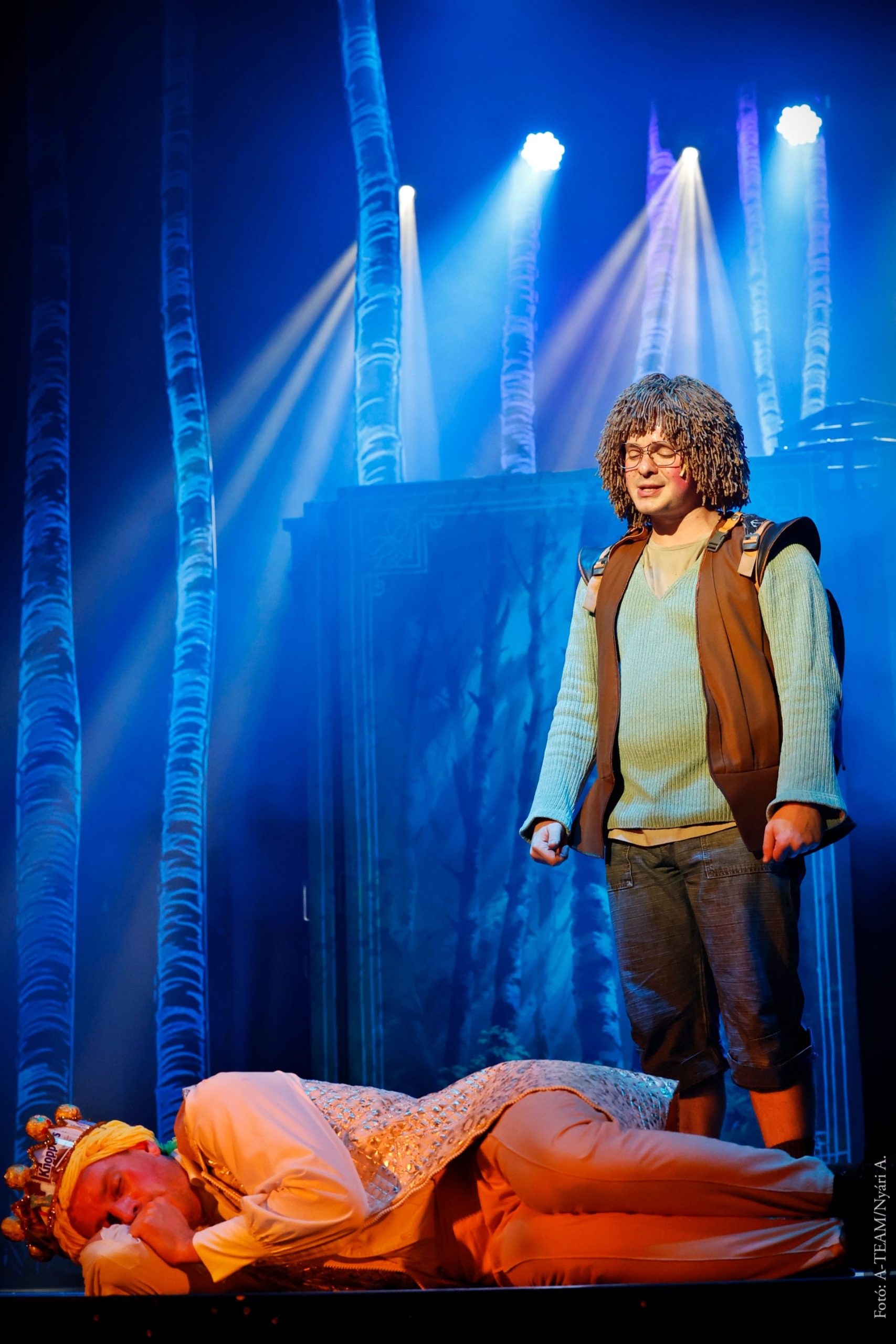 Színpadon egy férfi áll kócos parókában, mellette a földön fekszik egy koronás szereplő. A háttérben kékes fényben megvilágított erdei díszlet látható.