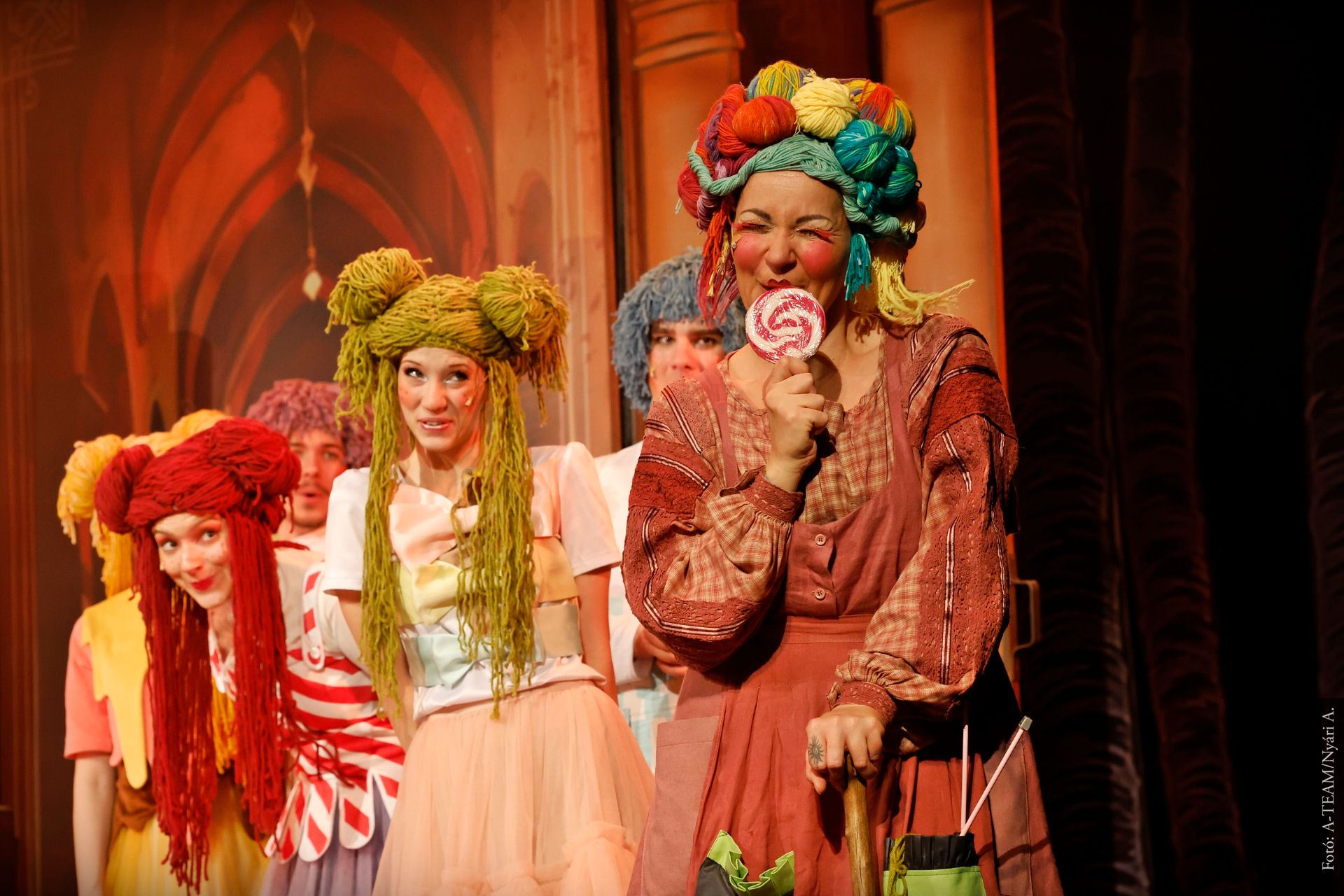 Színpadon több színész vidám jelenetben. Elöl egy nő áll színes ruhában és parókában, kezében nagy nyalókát tart, mögötte a többi szereplő mosolyog és figyeli.