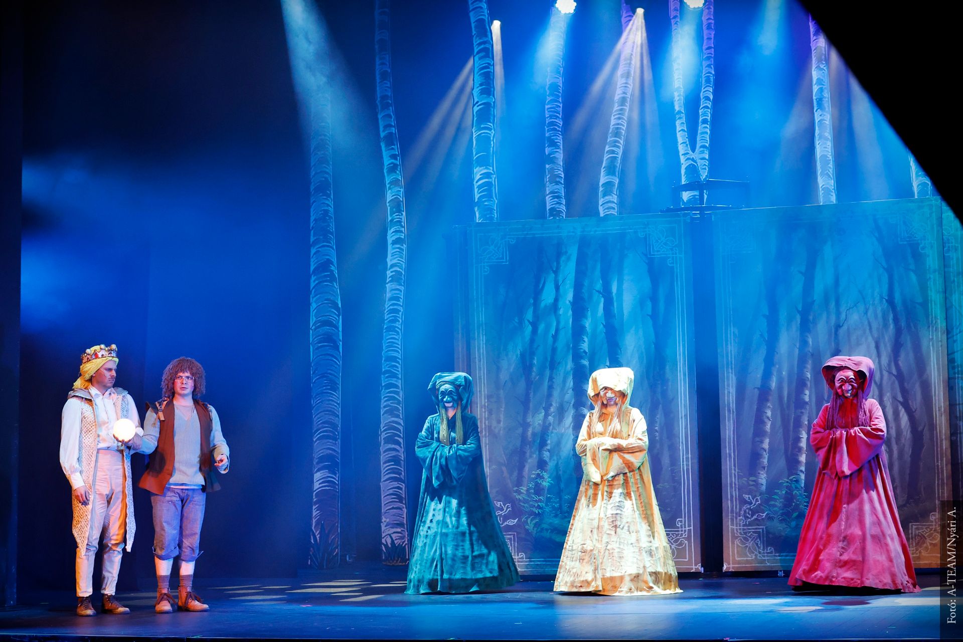 Színpadon öt színész áll erdei díszlet előtt, kékes fényben. Bal oldalon egy koronás férfi világító gömböt tart, mellette egy másik férfi, jobb oldalon három hosszú ruhás alak különböző színekben – kék, sárga és piros – figyelik őket.