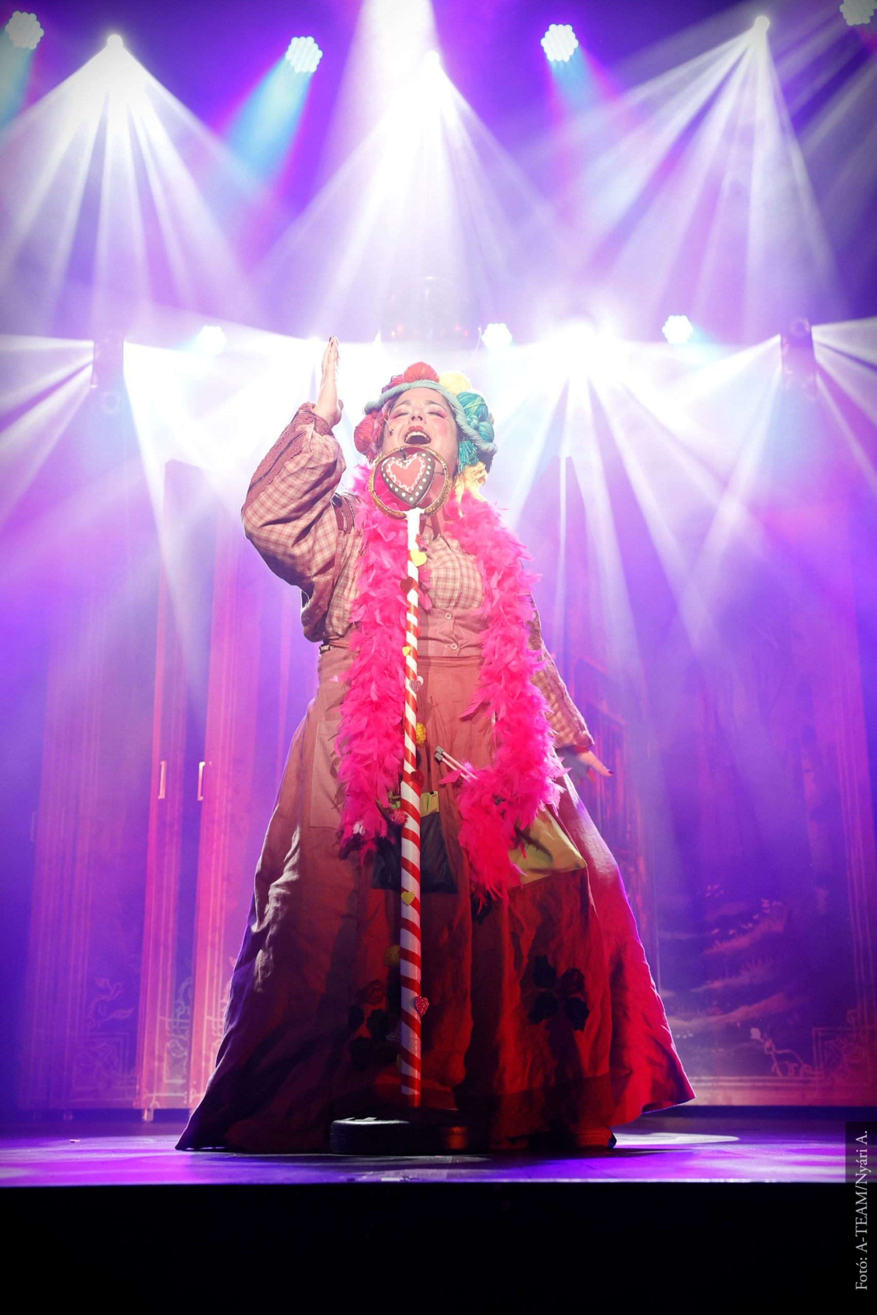 Színpadon álló színész színes, díszes jelmezben és parókában, mikrofon előtt énekel, mögötte látványos színes reflektorok fényei világítanak.