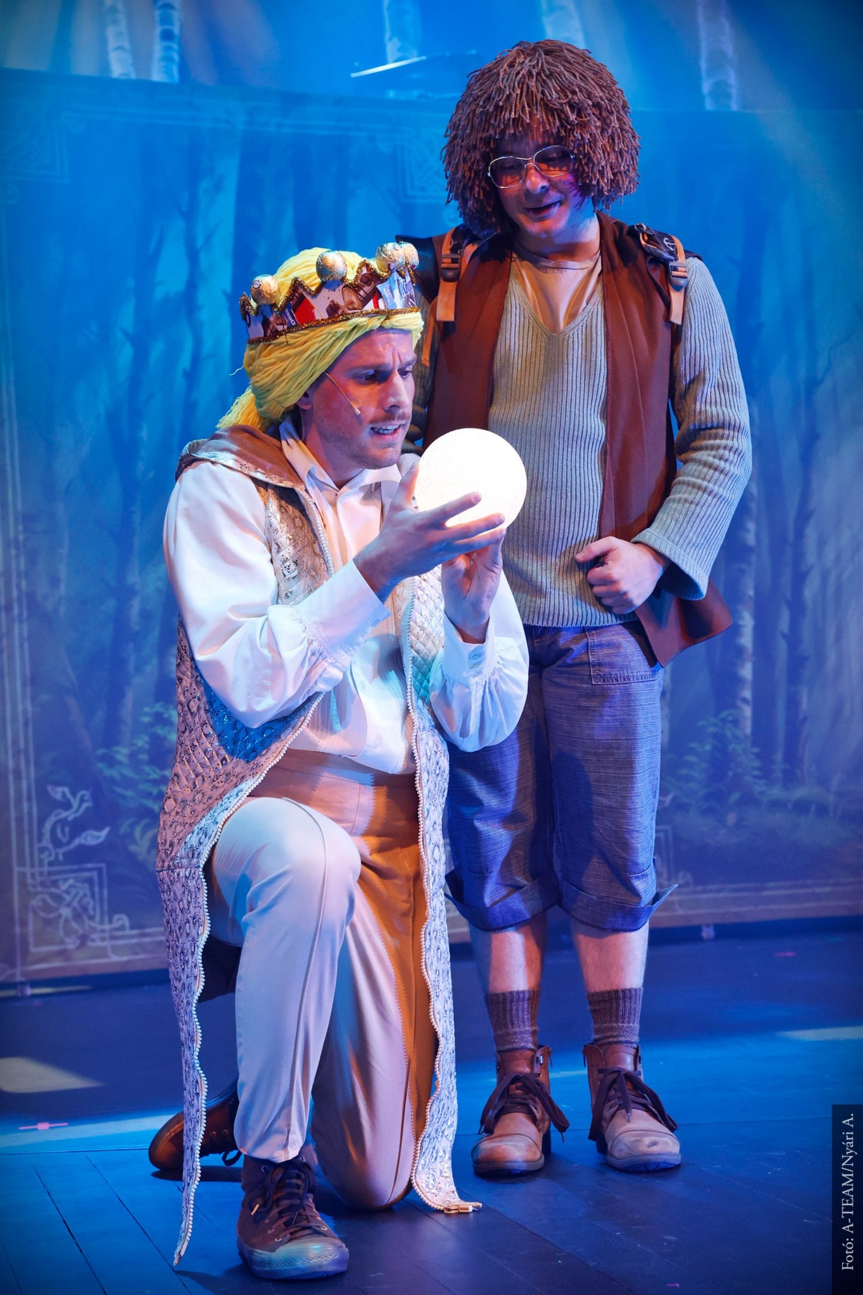 Egy koronás férfi térdel a színpadon, kezében világító gömböt tart, miközben egy másik, göndör parókás férfi áll mögötte, figyelmesen nézve. A háttérben erdei díszlet látható.