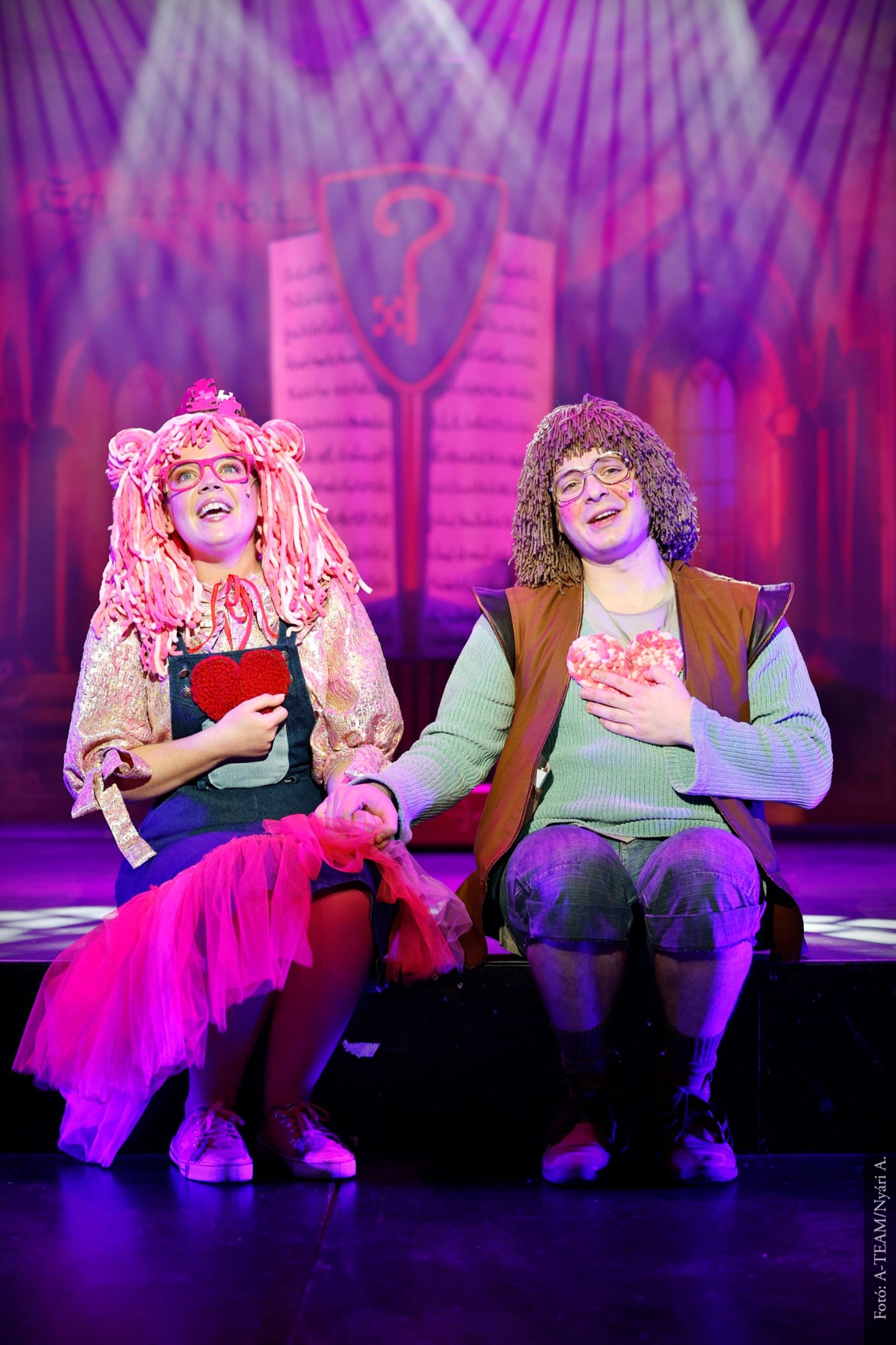 Két színész ül egymás mellett a színpadon, vidáman énekelnek vagy beszélnek, kezükben szívet formázó kellék. A háttérben színes fények és könyv alakú díszlet látható.
