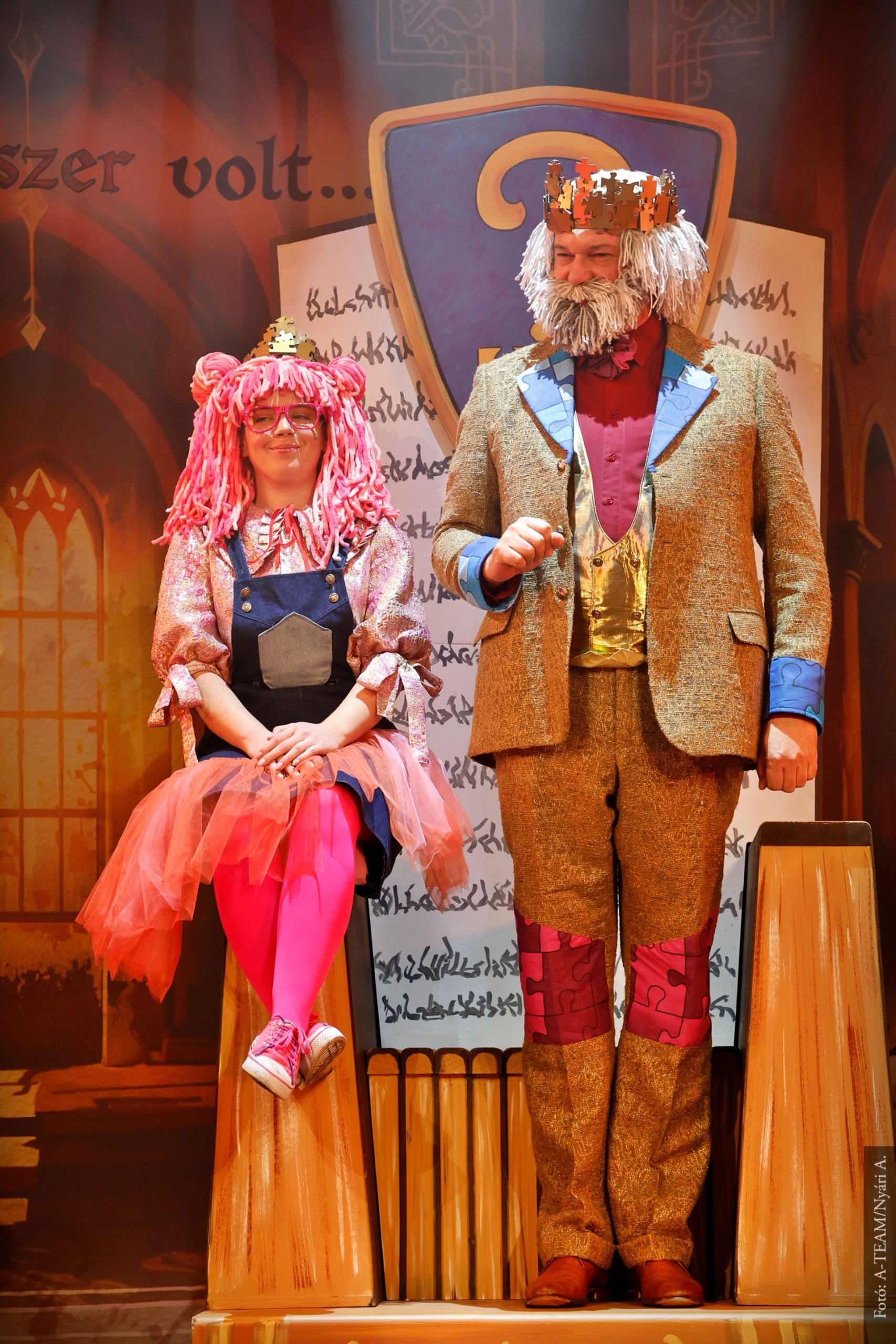 Színpadon egy koronás férfi és egy rózsaszín parókás nő ülnek és állnak egymás mellett, színes díszlet előtt. A férfi elegáns, arany részletekkel díszített ruhát visel, a nő tüllszoknyás jelmezben mosolyog.