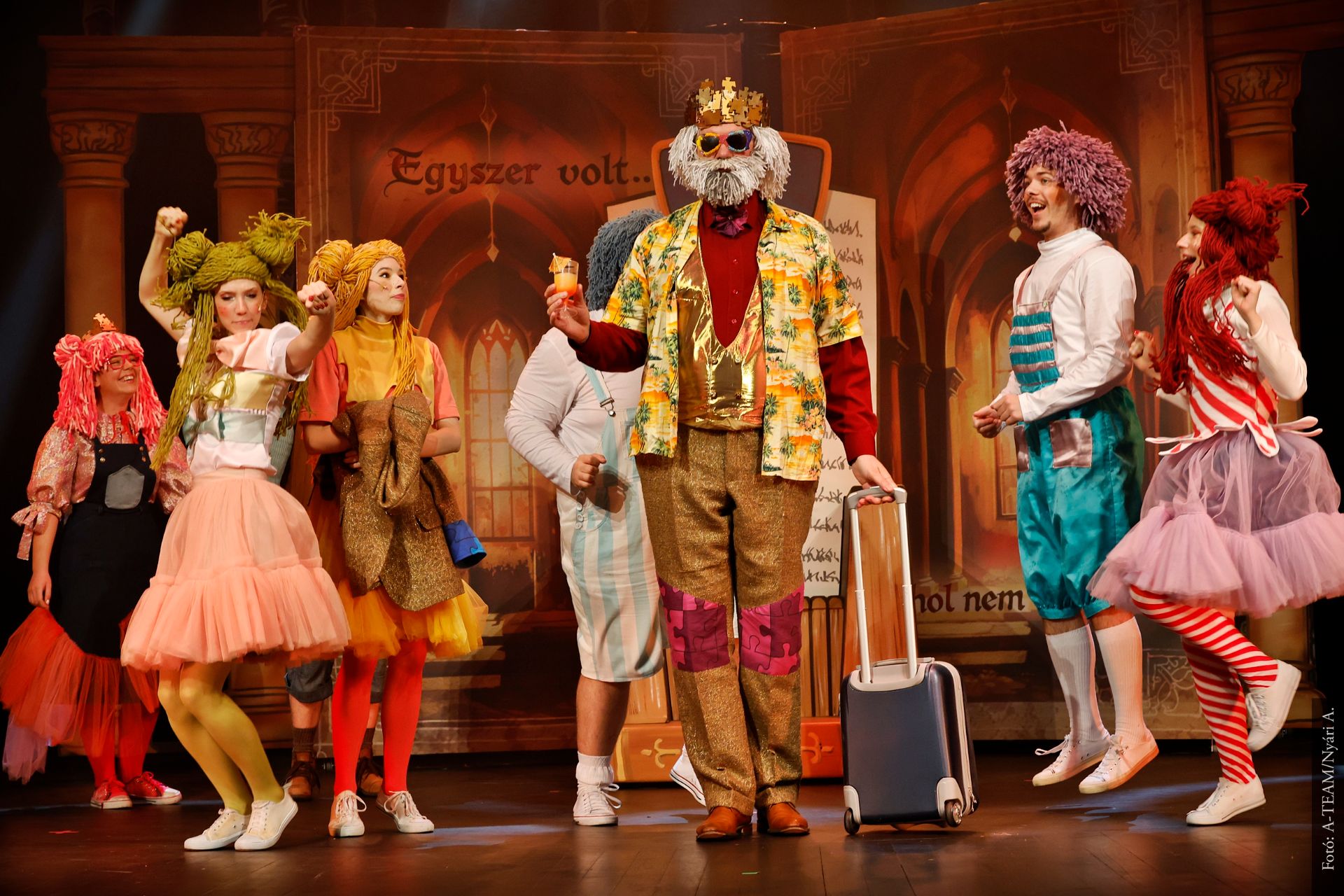 Színes jelmezes színészek állnak a színpadon vidám jelenetben. Középen egy koronás, napszemüveges férfi áll bőrönddel és italt tart a kezében, körülötte táncoló és nevető szereplők.
