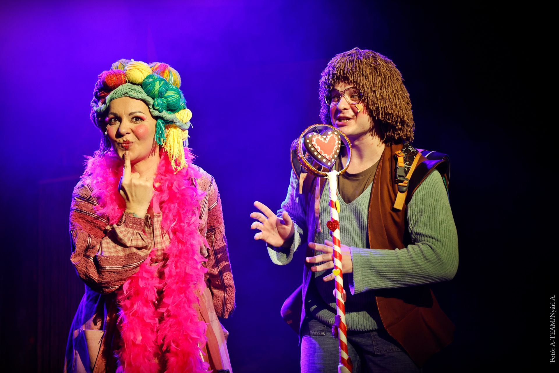 Két színész áll a színpadon színes jelmezben, vidám hangulatú jelenetben. A nő élénk rózsaszín tollboát visel, a férfi szív alakú díszítésű mikrofont tart a kezében.