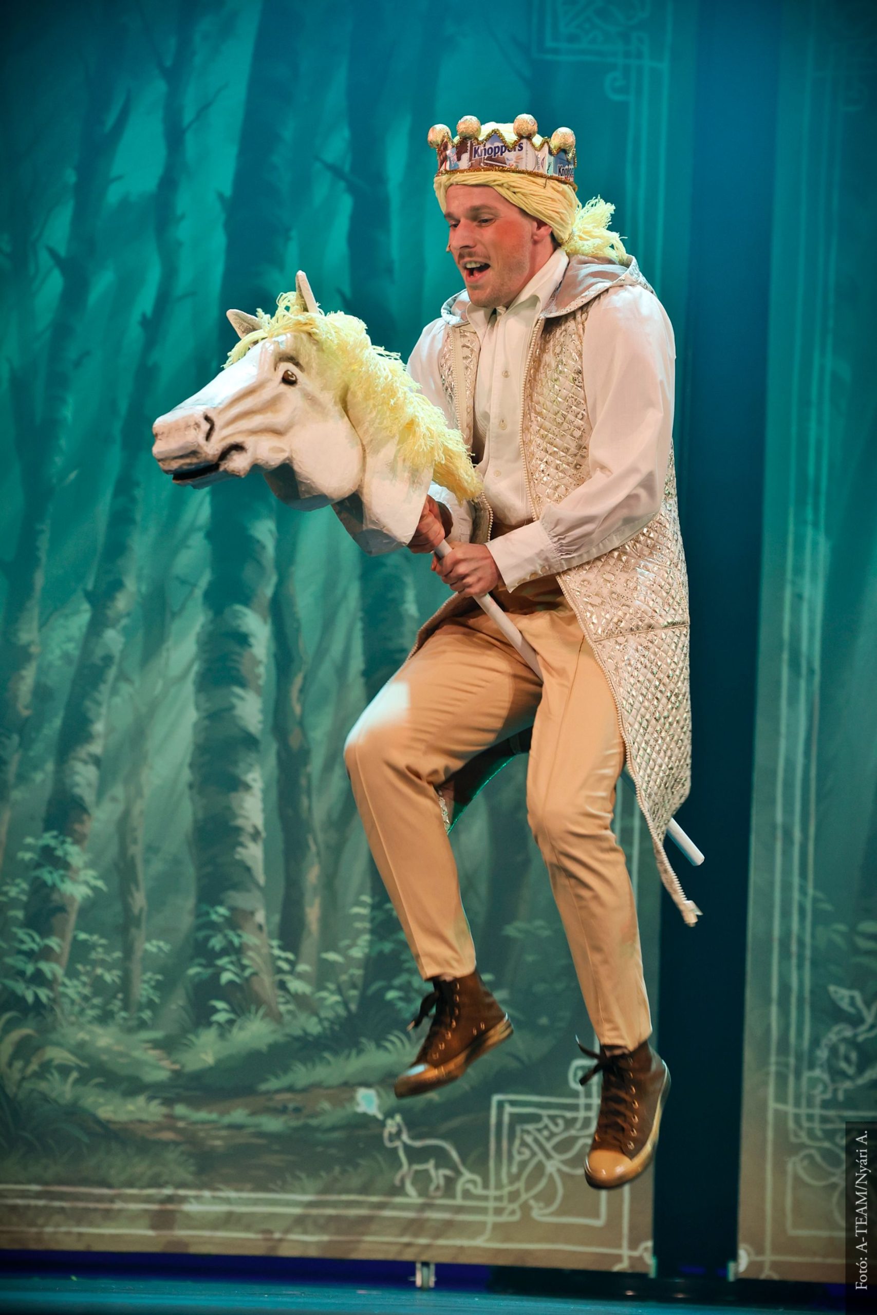 Koronás férfi jelmezben a színpadon, játék lófejet tartva lovaglást imitál. A háttérben erdei díszlet látható, a jelenet vidám hangulatú.