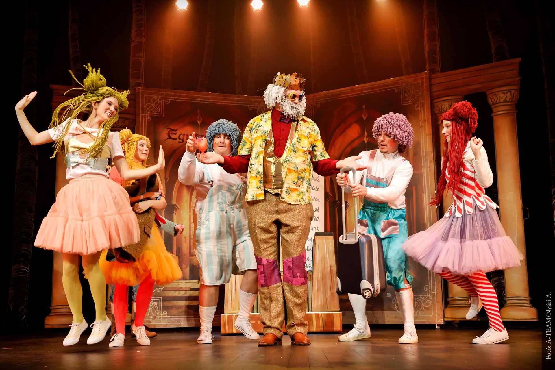 Színes jelmezes színészek egy vidám színházi jelenetben a színpadon. Középen egy koronás, napszemüveges férfi áll bőrönddel és itallal a kezében, körülötte táncoló szereplők élénk ruhákban.