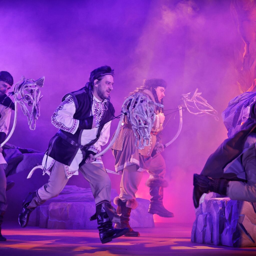 Férfiak magyar népi ruhában futnak, a kezükben egy csontváz ló fej, a háttérben füst van