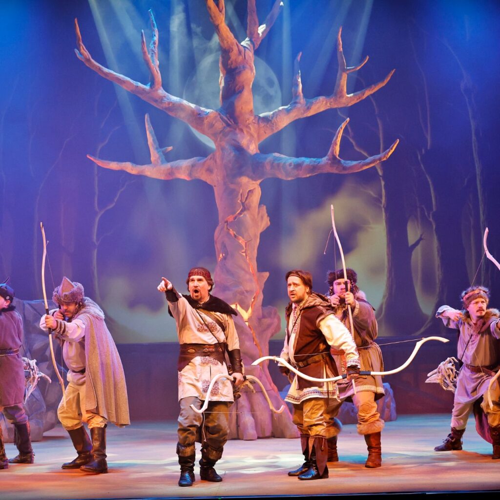 Hat férfi ősmagyar ruhában áll a színpadon, a kezükben íj van. Középen aki áll a távolba mutat és kiabál. Mögöttük egy díszlet fa van