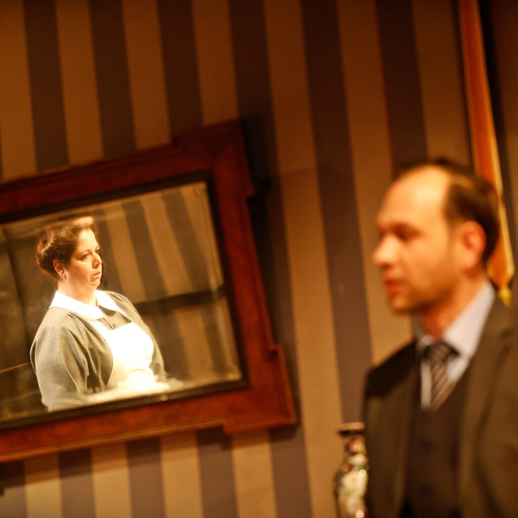 Bal oldalt egy tükör látható a képen, ami a falra van akasztva, és egy fehér kötényes nő látható benne. Jobbról egy férfi alakja jelenik meg elmosódva