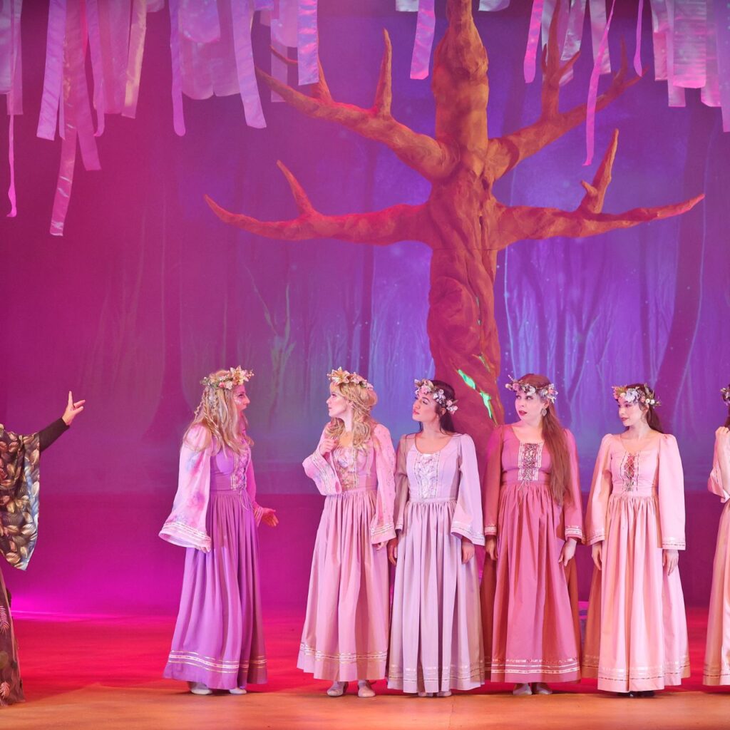 Hét nő áll a színpadon, a jobb oldali hat személy rózsaszín tündérruhában van és beszélgetnek, a bal oldalt álló nő pedig tündérkirálynőnek van öltözve, zöld virágos ruhája van és éppen beszél. A háttérben egy nagy díszlet fa van