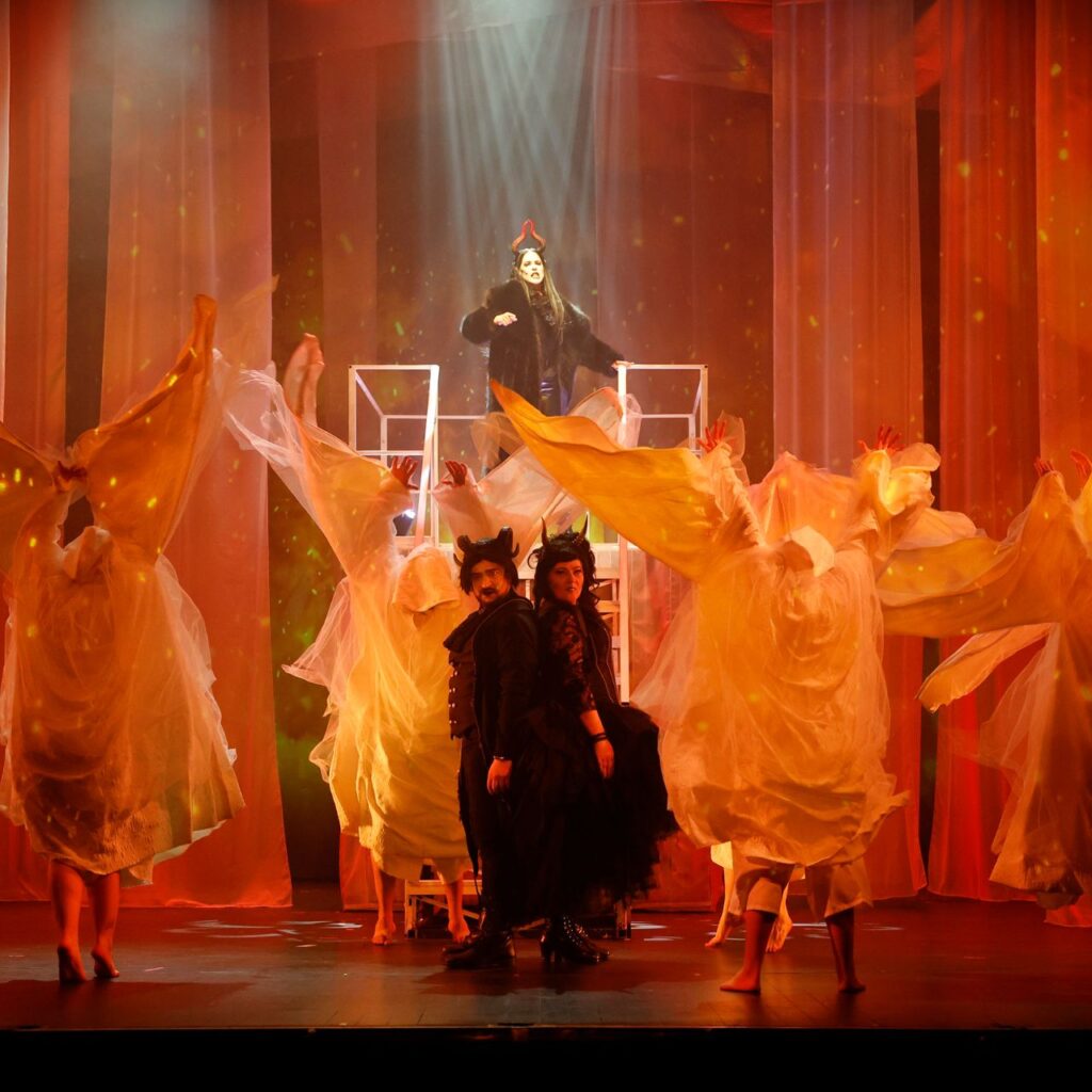 Fekete ruhás ördögnek öltözött emberek állnak a kép közepén hárman, körülöttük fehér lepel ruhás emberek táncolnak. A fények vörösek