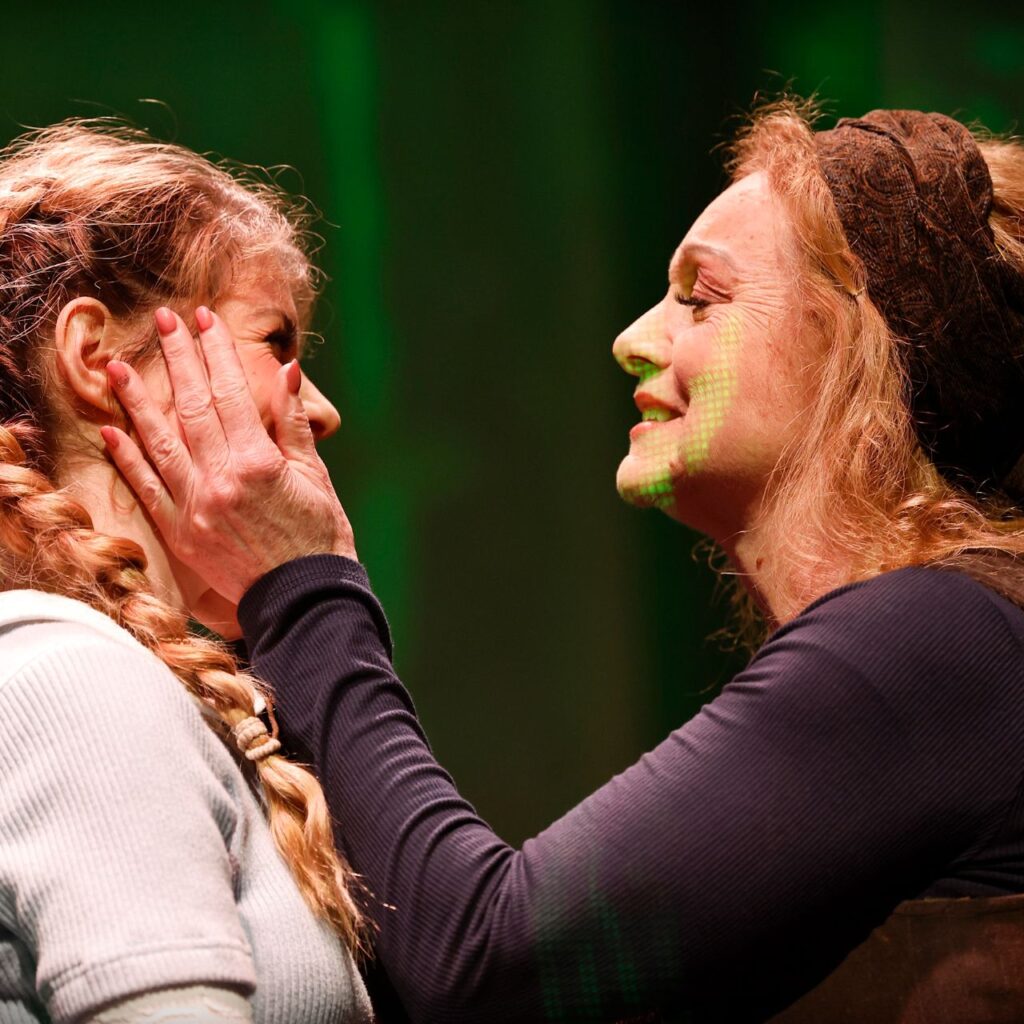 Két nő van a képen közelebbről fotózva, a bal oldali fiatalabb, a haja copfban van és nagyon mosolyog. A jobb oldali idősebb nő olyan mint az anyukája, a kezeivel a lány arcát fogja és büszkén néz rá. A háttér zöld fényekkel van megvilágítva