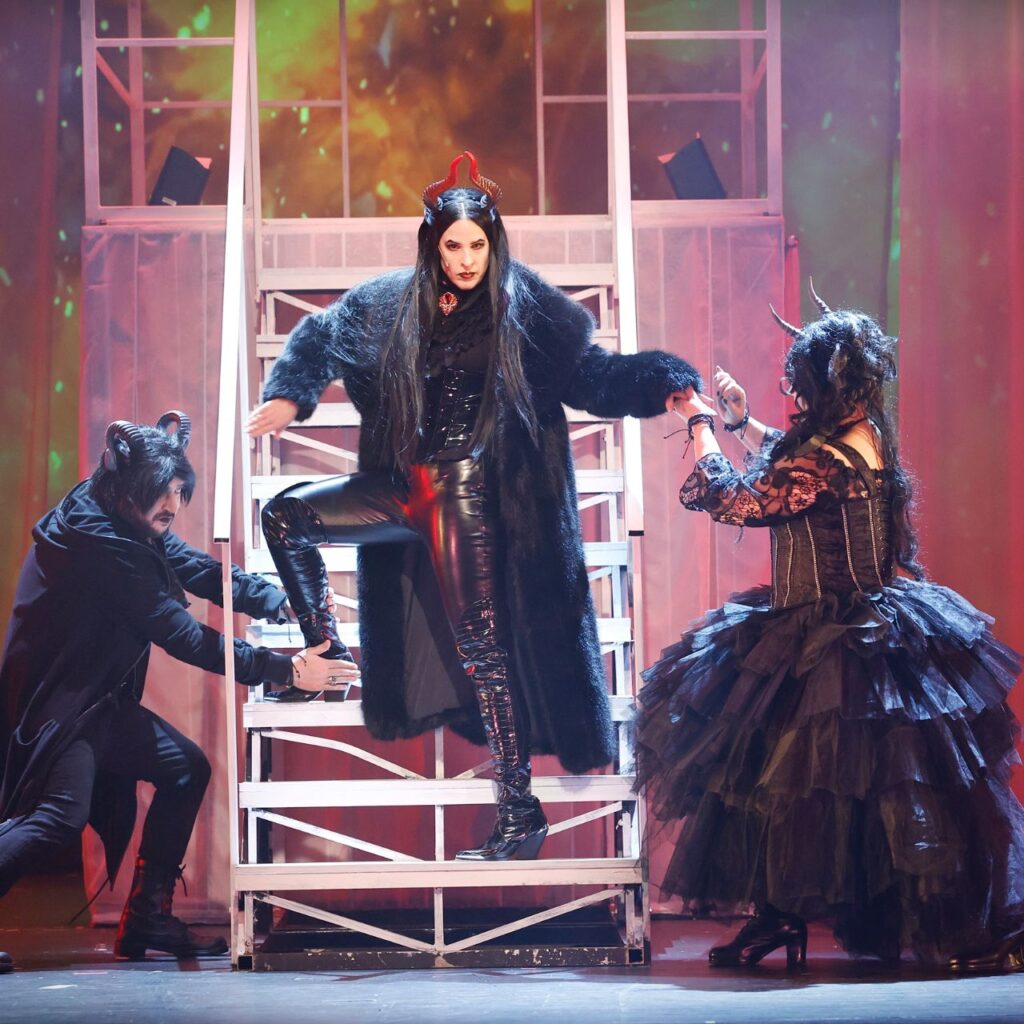 Vörös háttér előtt középen van egy lépcső, amiről egy ördögnek öltözött nő lépked le magabiztosan. A két kezénél fogva segíti le két oldalt szintén ördögnek öltözött nő és férfi