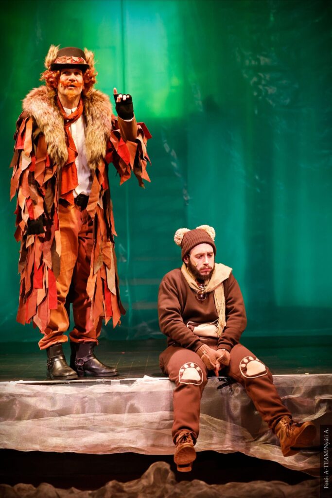 Egy zöld színű háttér előtt balról áll egy férfi, akinek az egész ruhája vörös levél szerű textilből áll, vörös nagy bajsza van kalapja. Feltartja az egyik kezét és éppen úgy beszél. Mellette a kép jobb oldalán egy medvének öltözött férfi ül a színpad szélén, és szomorúan néz maga elé.