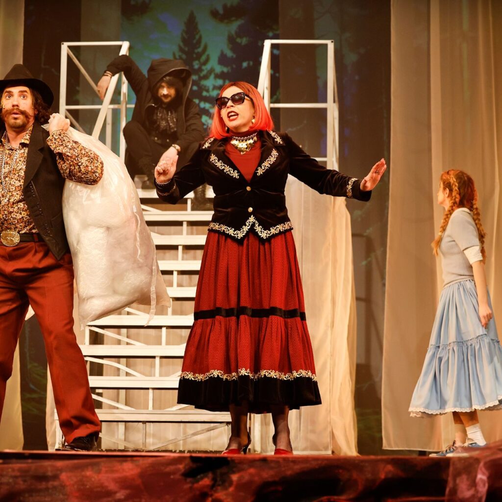 A kép közepén egy nő áll piros szoknyában és fekete blézerben, oldalra mutogatva beszél, a fején napszemüveg van és rózsaszín a haja. Mellette balról egy kalapos és mellényes férfi áll, akinek két nagy zsák van a kezében. A háttérben még további három személy látható, akiknek nem látszódik az arca mert hátrafele néznek