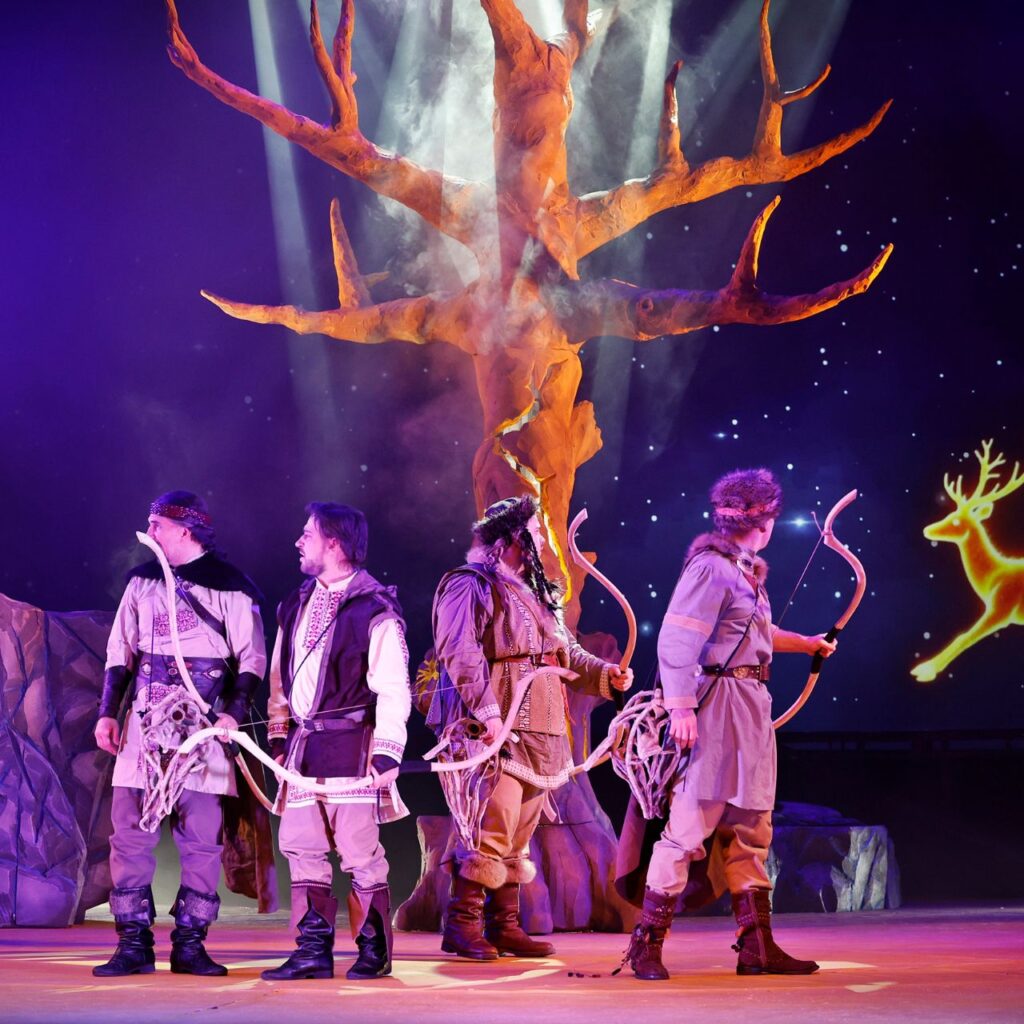 A képen öt férfi látható ősmagyar ruhában, a kezükben íjak vannak, és jobb oldalra néznek, ahol egy falra vetített szarvas fénylik a csillagos égen. A háttérben még látható egy nagy díszlet fa