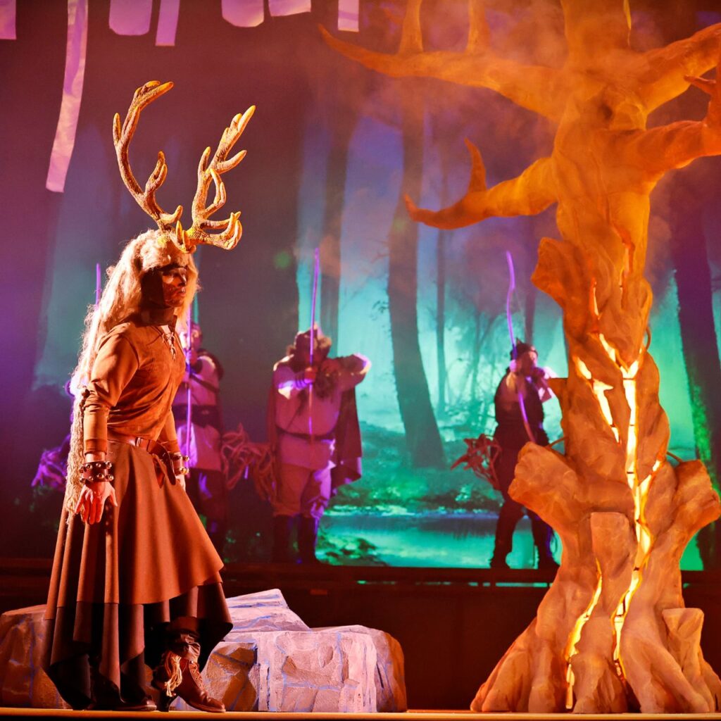 A képen bal oldalt egy szarvasnak öltözött férfi sétál, jobb oldalt egy díszlet fa van, a háttérben pedig halványan látható öt férfi, akik a szarvasra szegezik az íjaikat.