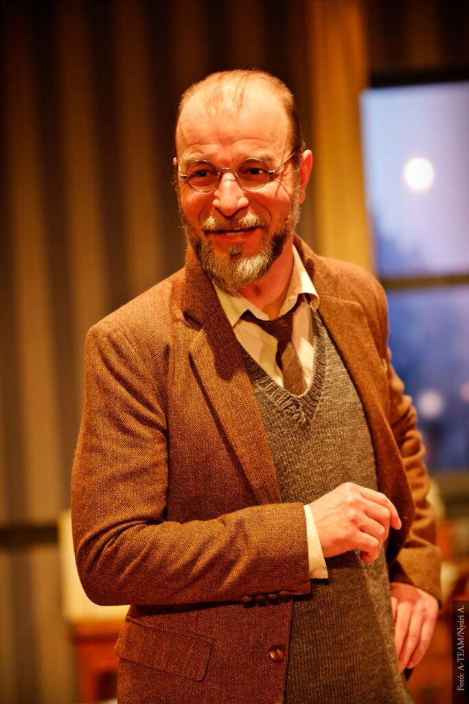 Egy férfi áll a képen, éppen mosolyog. Kerek szemüveget visel, barna zakóban van, alatta egy szürke kötött mellány, ami alól kilátszódik egy nyakkendő és a fehér ing.