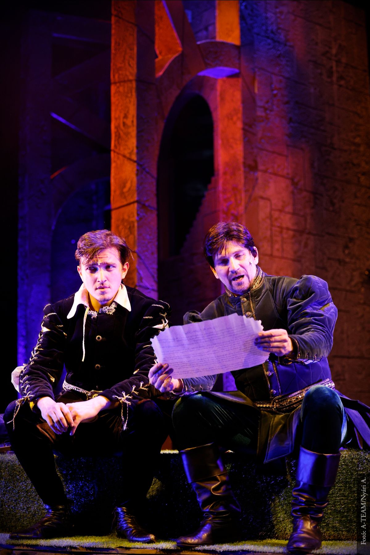 A színművészek a Hamletrock című előadásban jelenet közben a színpadon a díszletfal előtt ülve párbeszédet folytatnak.