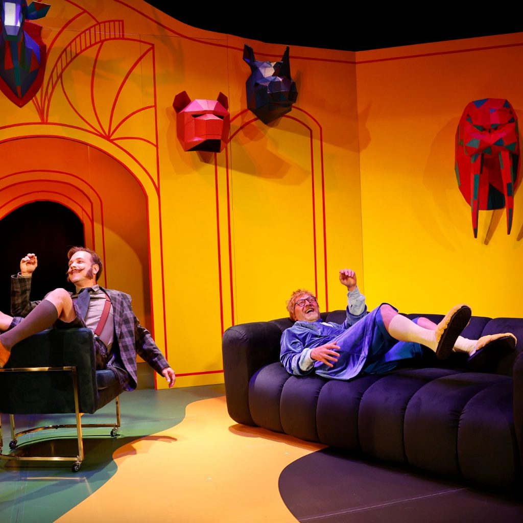 A Kék róka című színházi előadás vidám jelenete: két férfi nevetve ül és fekszik a színes, sárga díszletű szobában.