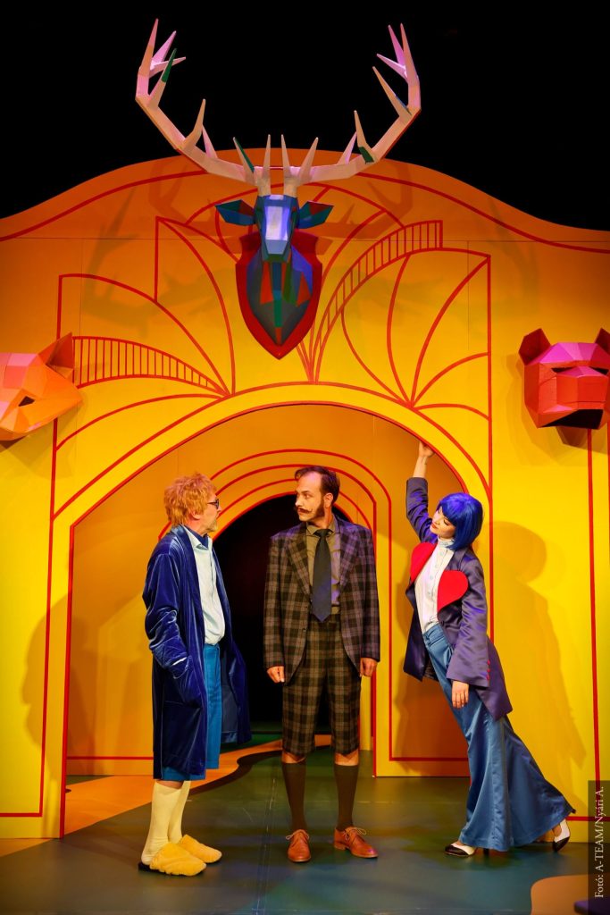 A Kék róka című színházi előadás egyik jelenete: három szereplő a színes, sárga díszlet előtt áll, felettük egy stilizált agancsos szobor látható.