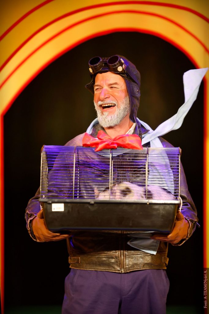 A Kék róka című színházi előadás egyik jelenete: egy mosolygó férfi pilótasapkában, kezében egy kalitkával, benne egy kisállattal.