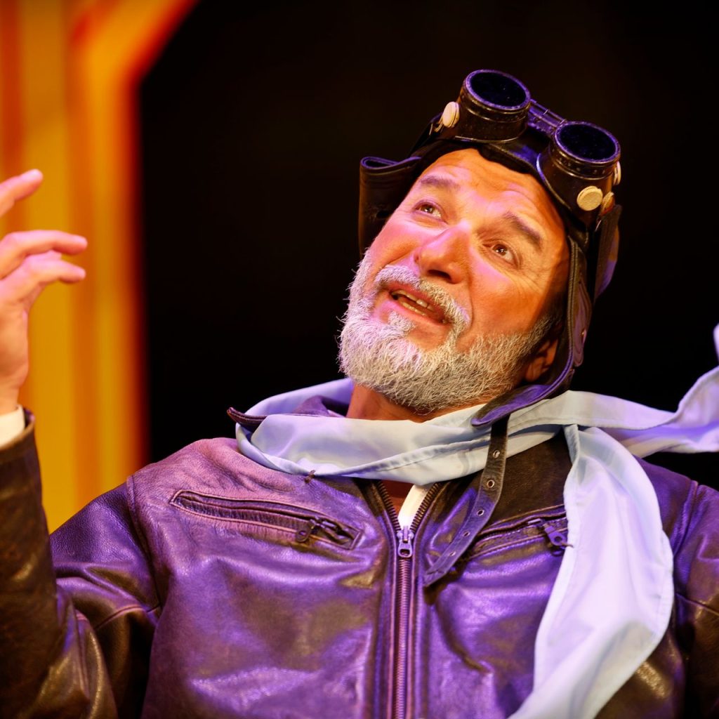 A Kék róka című színházi előadás egyik jelenete, egy férfi pilótasapkában és sálban, felemelt kézzel beszél a színes díszlet előtt