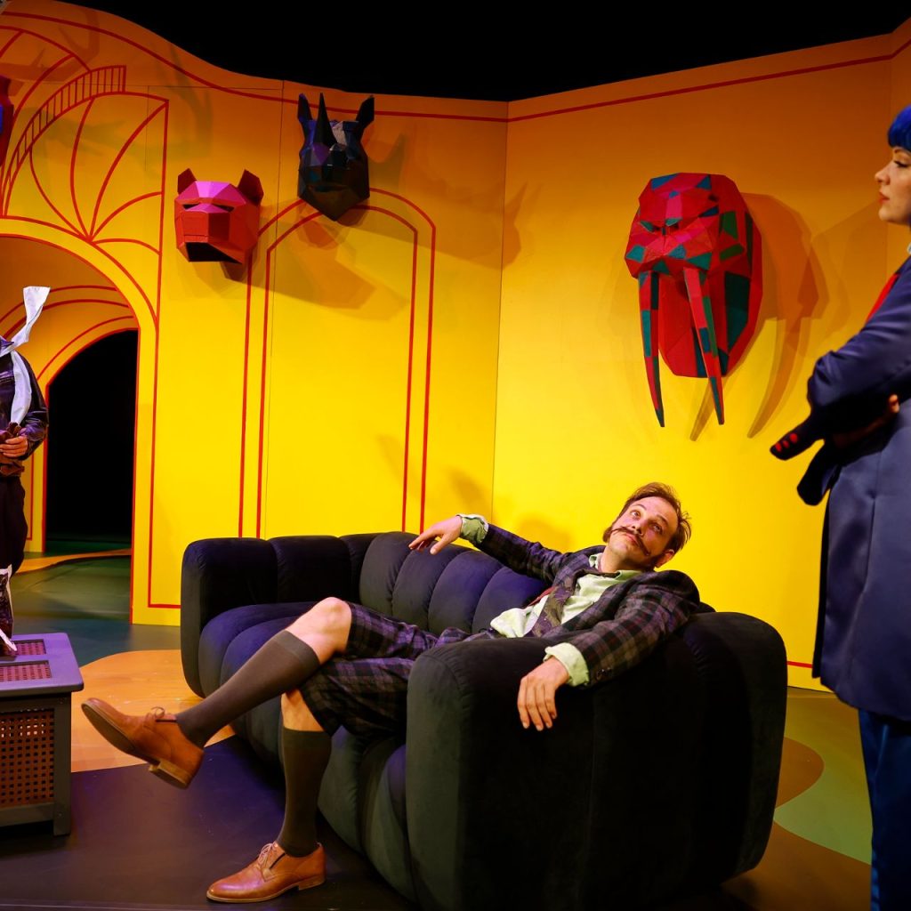 A Kék róka című színházi előadás egyik jelenete: három szereplő színes, sárga díszletben, egy férfi a kanapén, egy másik áll, a nő kék parókában velük szemben.