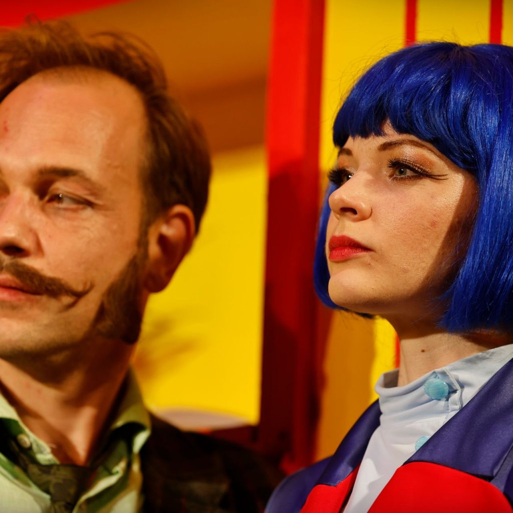 A Kék róka című színházi előadás két szereplője közelről, színes díszlet előtt – a férfi zöld zakóban, a nő kék parókában és lila kabátban.