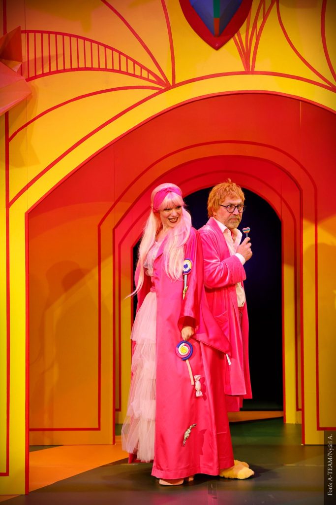 A Kék róka című színházi előadás két szereplője élénk rózsaszín jelmezben, színes díszlet előtt áll a színpadon.”