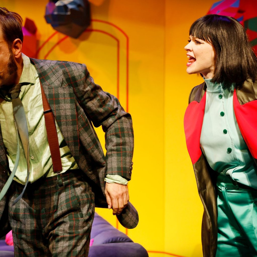 A Kék róka című színházi előadás jelenete, két szereplő heves párbeszéd közben a színpadon, színes díszlet előtt.