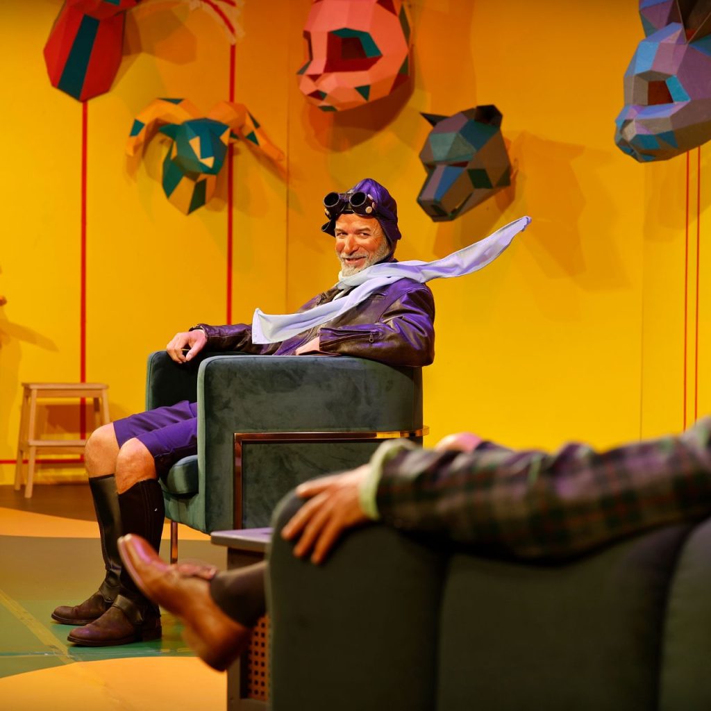 Egy férfi vintage pilótaöltözetben, védőszemüvegben és fehér sállal ül mosolyogva egy zöld széken egy élénksárga, színes állatfejekkel díszített szobában. Egy másik személy az előtérben ül, háttal a kamerának.