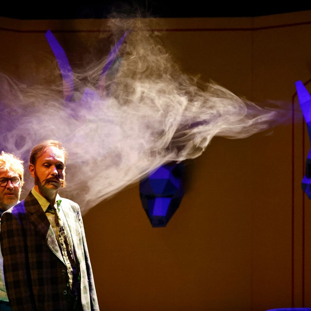 Két öltönyös férfi áll a színpadon drámai megvilágításban, fölöttük füst kavarog. Mögöttük kék, geometrikus állatfejek vannak egy sárga falra szerelve, szürreális és színházi hangulatot teremtve.