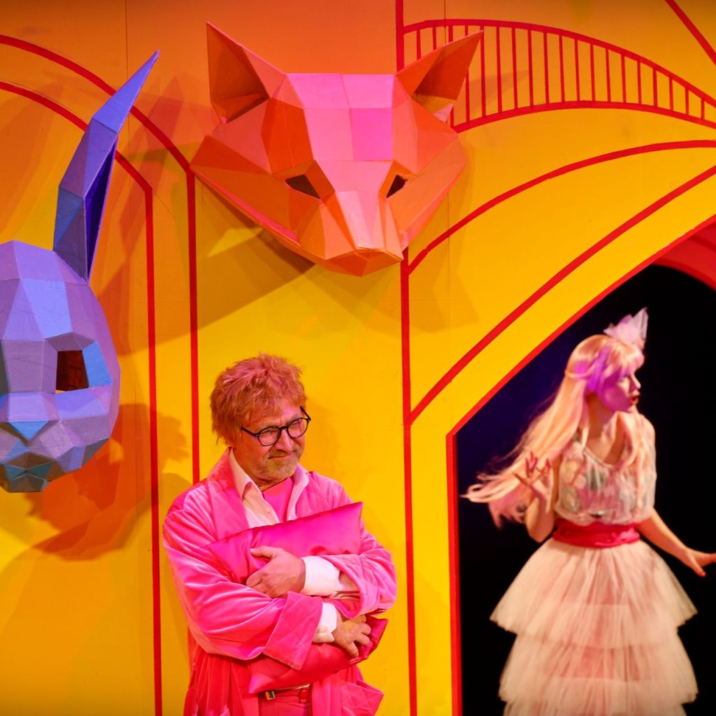 Egy rózsaszín köpenyes férfi áll keresztbe tett karokkal, nagy, színes, geometrikus állatmaszkok alatt egy élénksárga színpadon. Egy nő fodros ruhában és állatmaszkban áll az ajtóban, és a férfi felé néz.