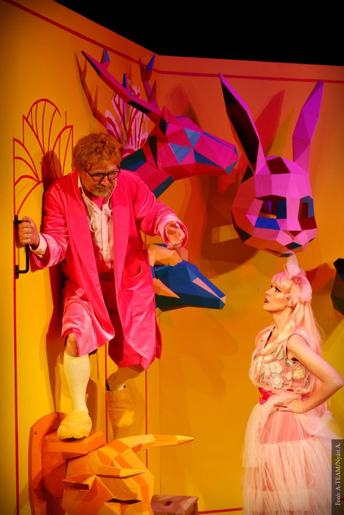 Egy élénk rózsaszín öltönyös, zoknis férfi áll narancssárga állatos kellékeken, és egy rózsaszín ruhás, fejdíszes nővel beszélget. Színes geometrikus állatfejek díszítik a sárga háttérfalat.