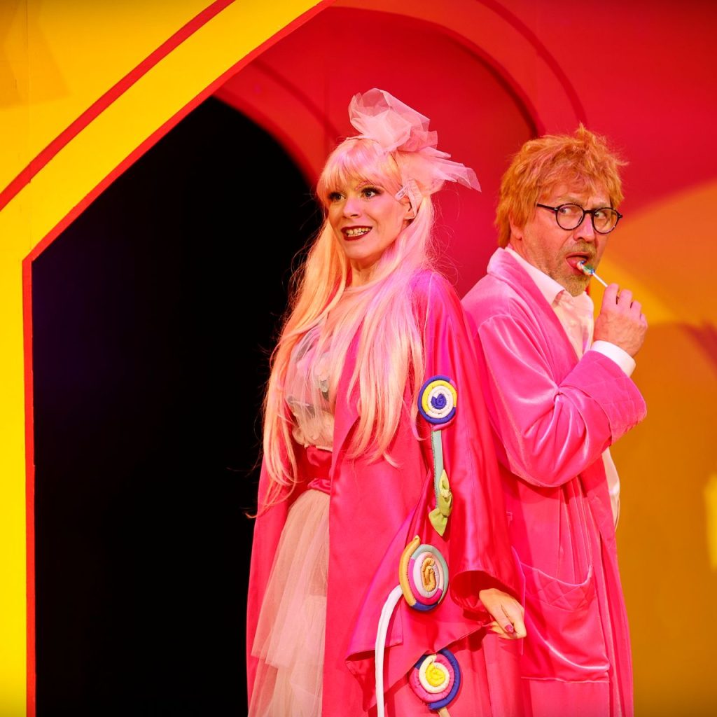 Két felnőtt élénk rózsaszín jelmezben áll háttal egymásnak. A nőnek hosszú rózsaszín haja, nagy masnija és nyalókadíszek vannak a köntösén, míg a férfi szemüveget és rózsaszín köpenyt visel, és egy botot rágcsál. Színes, játékos összeállításban állnak.