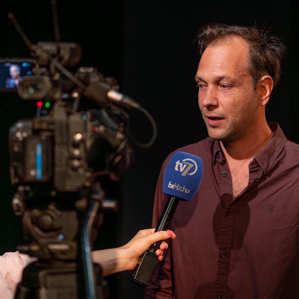 A Kék róka című előadás olvasópróbáján egy férfi interjút ad a TV7 csatornának, miközben egy kamera rögzíti a beszélgetést.