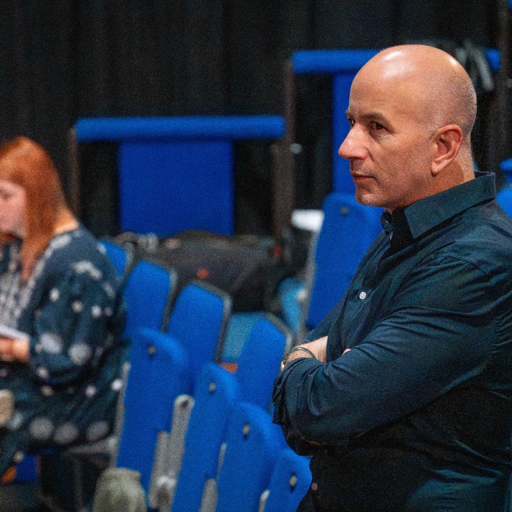 A Kék róka című színházi előadás olvasópróbáján a rendező figyeli a próbát, a háttérben a súgó szövegkönyvet tart.