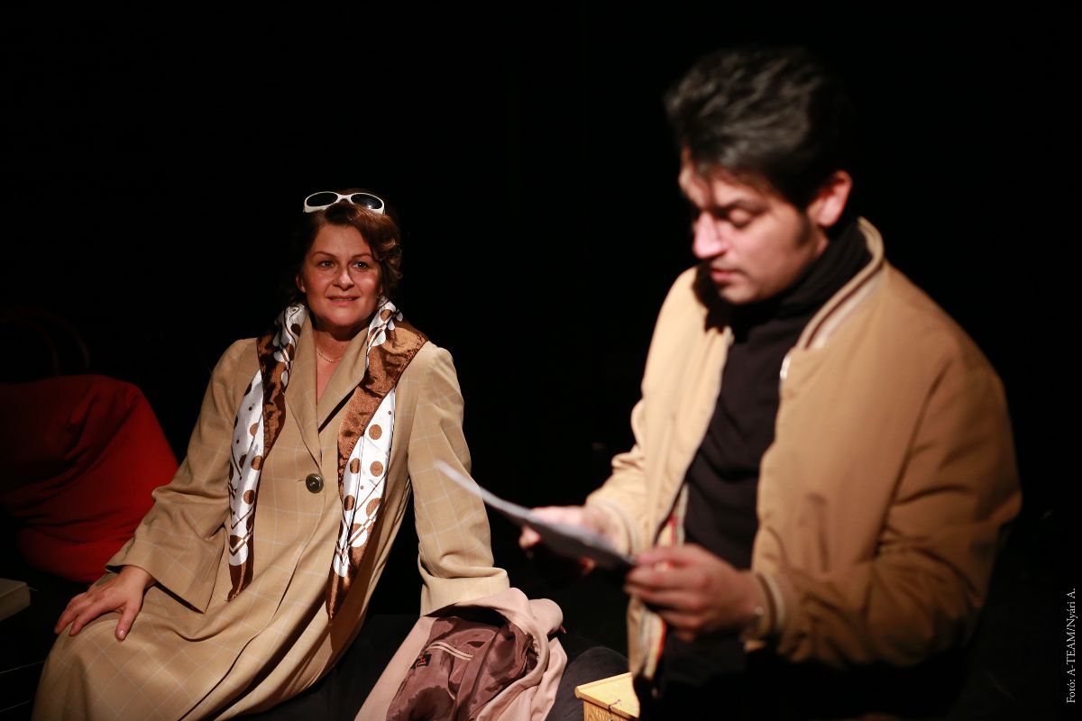 A színművészek a Két összeillő ember című előadásban jelenet közben a színpadon, a férfi olvas, a nő figyeli.