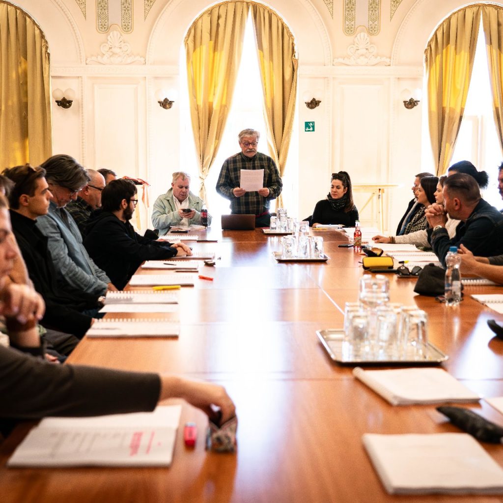 A Látni a láthatatlant című előadás olvasópróbáján a társulat egy hosszú asztal körül ül, az egyik szereplő állva olvassa a szöveget.